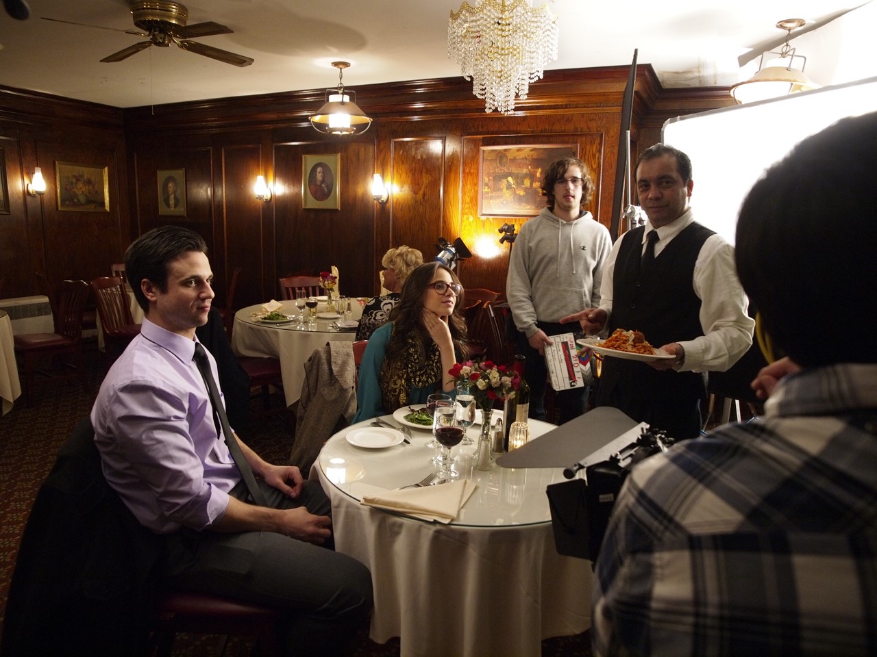 Actors in a restaurant scene with a waiter serving pasta, capturing a film or dinner setting.