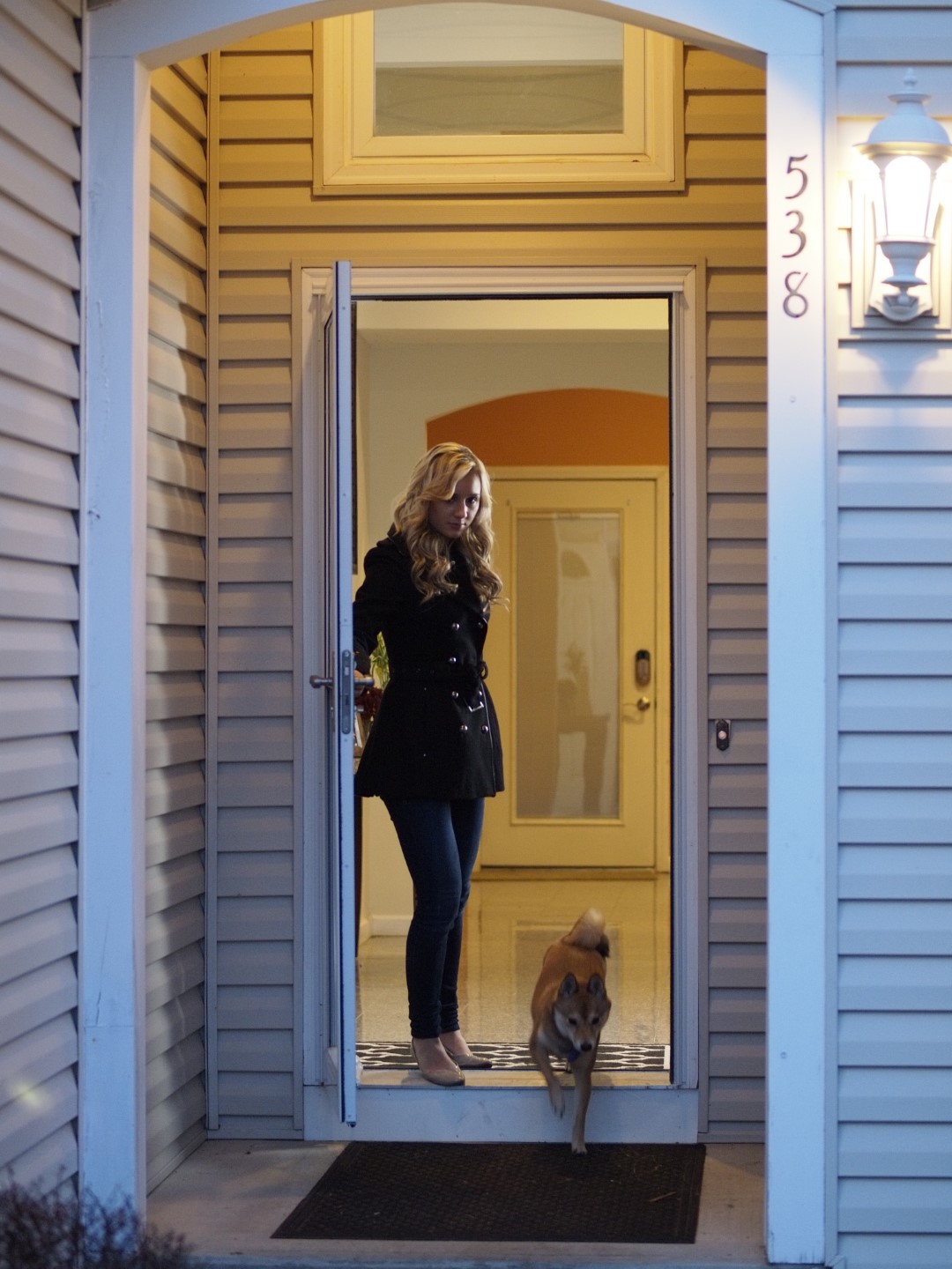 Julie Dawson in black coat standing at home entrance with a dog stepping out at dusk.