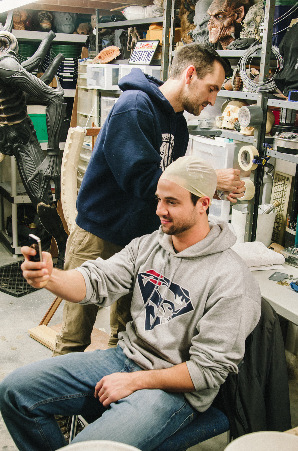 Two men in a workshop creating costumes, one using a smartphone and the other working on a headpiece.