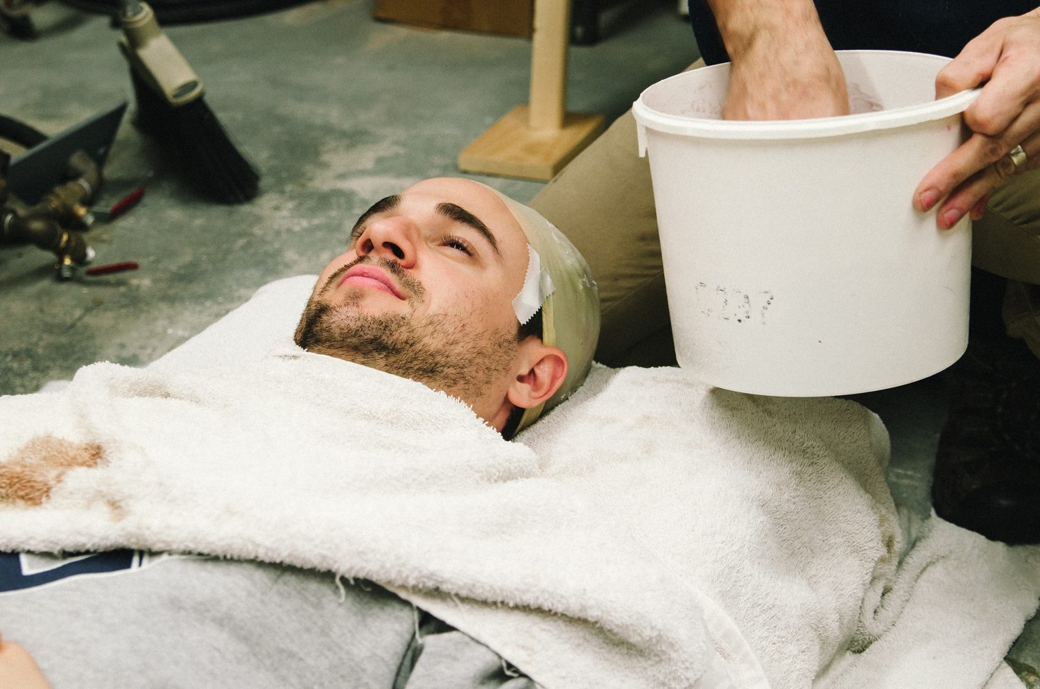A man lying on a towel, preparing for a process involving a bucket, in a workshop setting.