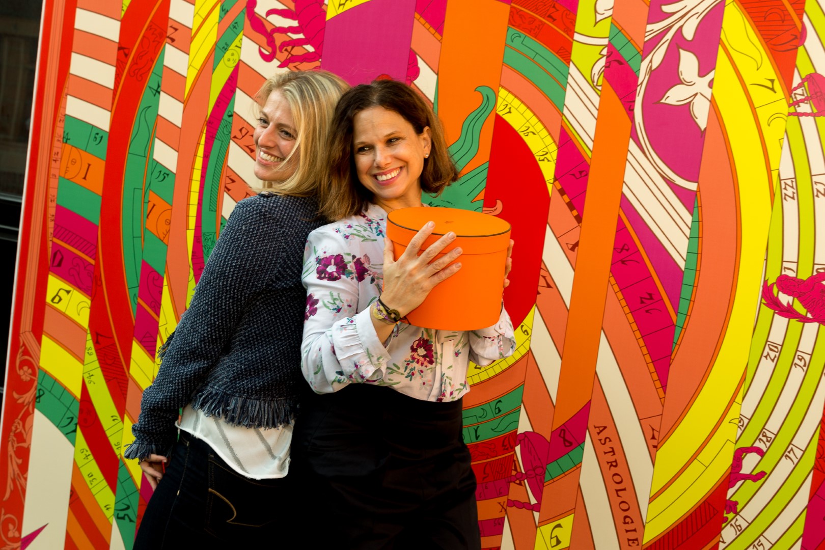 Two women smiling against a vibrant, colorful backdrop, one holding an orange box.