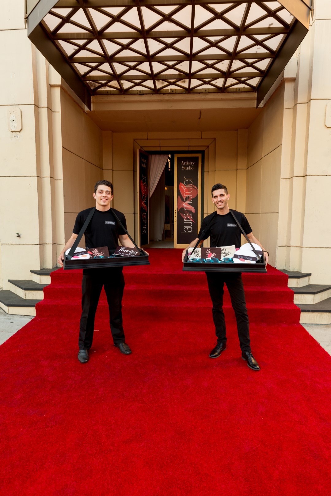 Two servers in black attire holding trays at a red carpet entrance to an artistry studio.