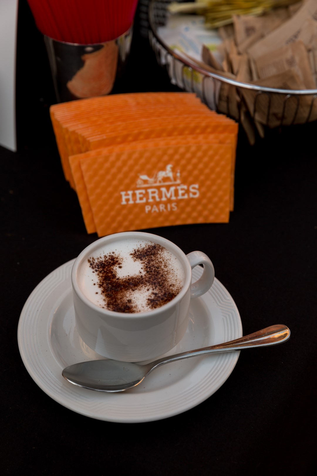 Cup of coffee with artistic foam design, alongside Hermes Paris sugar packets