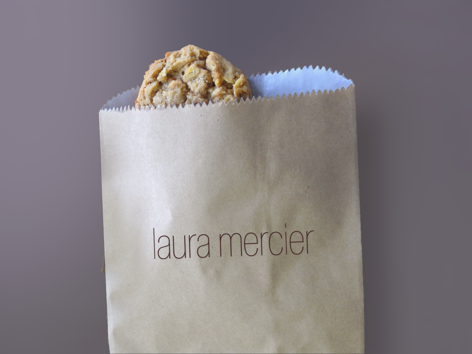 Oatmeal cookie in a branded laura mercier paper bag on a purple background.