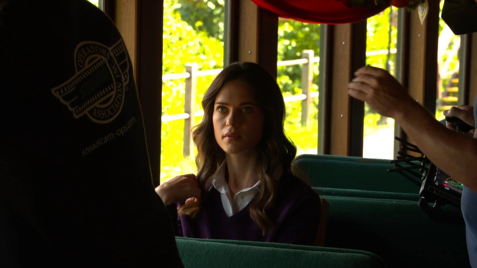 A young woman seated, looking attentive while a crew member operates camera equipment.