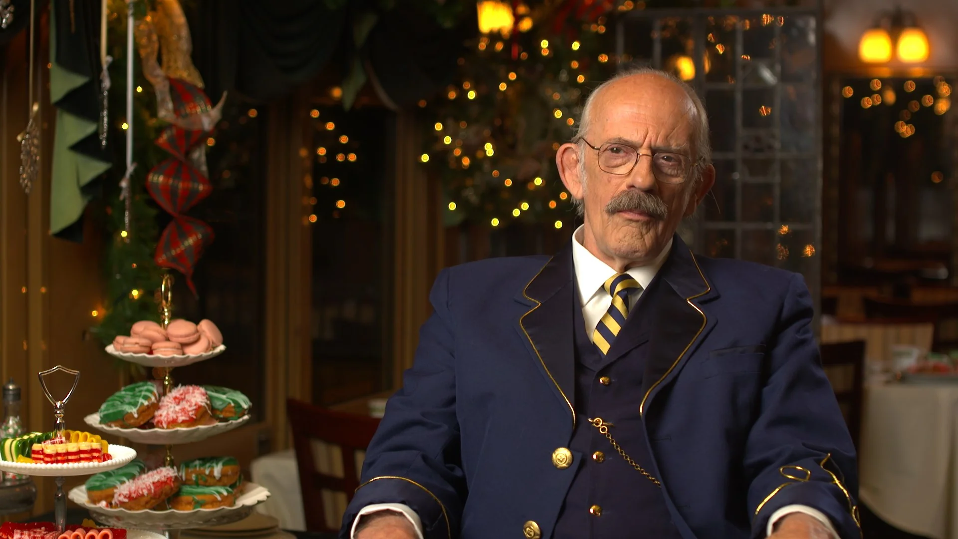 Christopher Lloyd in a formal suit sits by a dessert display in a festive setting.