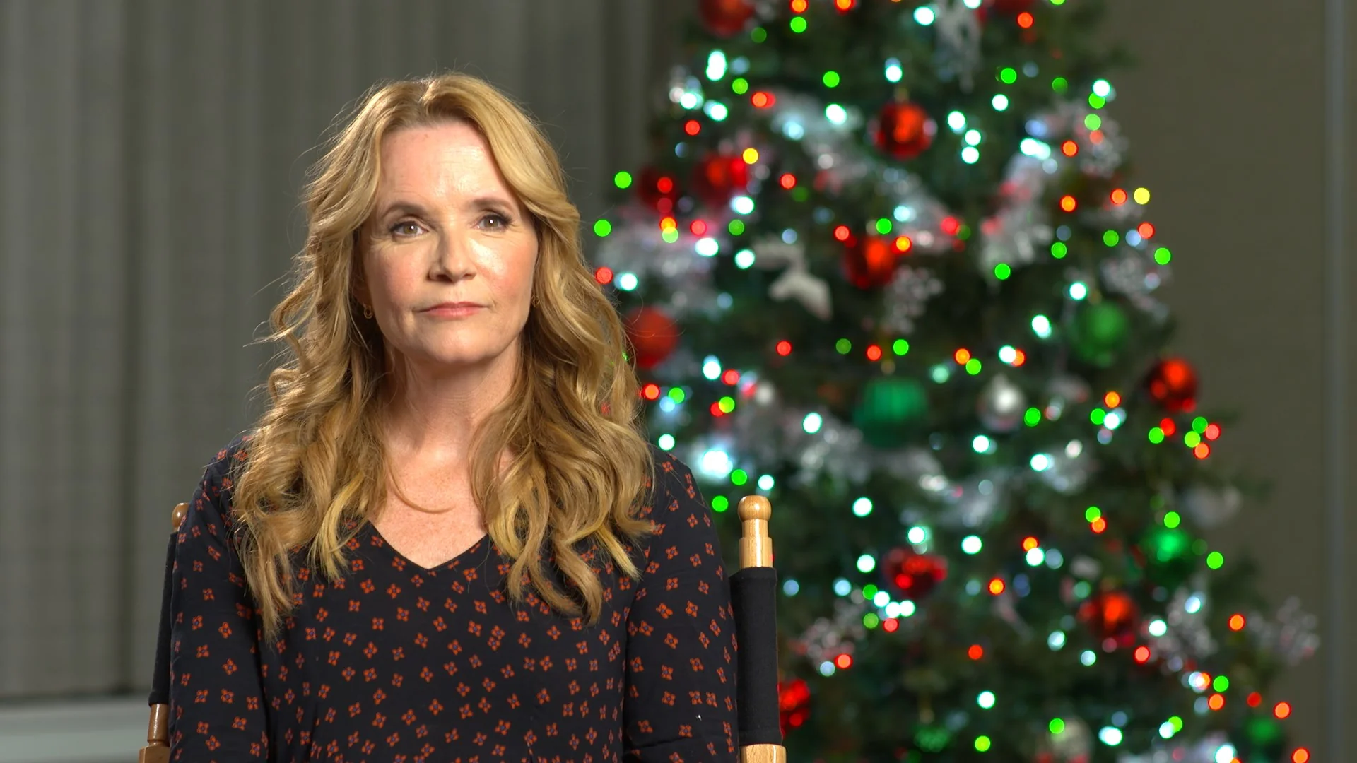 Lea Thompson sitting in front of a decorated Christmas tree with colorful lights.