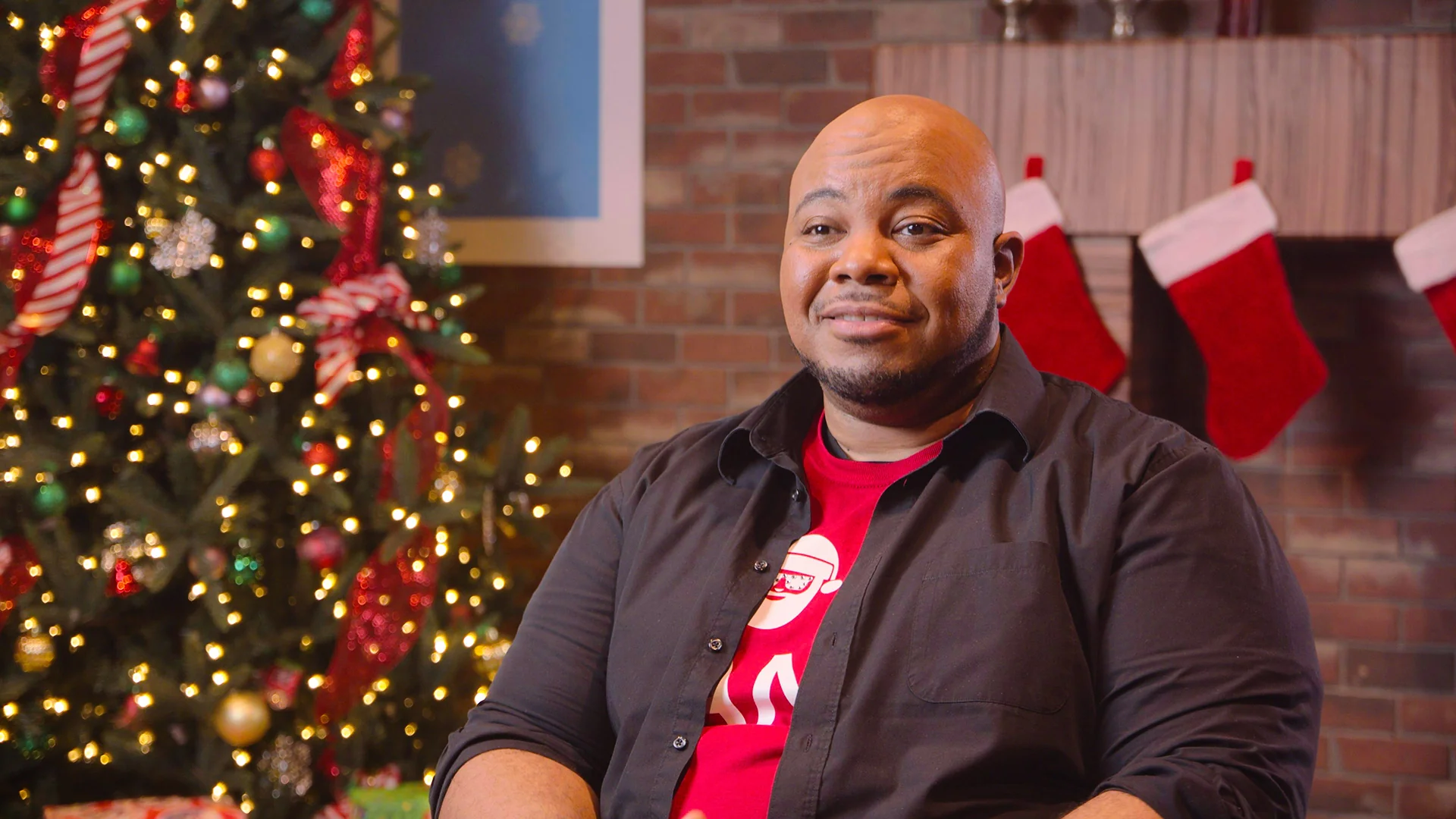 Man in a festive setting with Christmas tree and stockings, wearing a red shirt, smiling at the camera.
