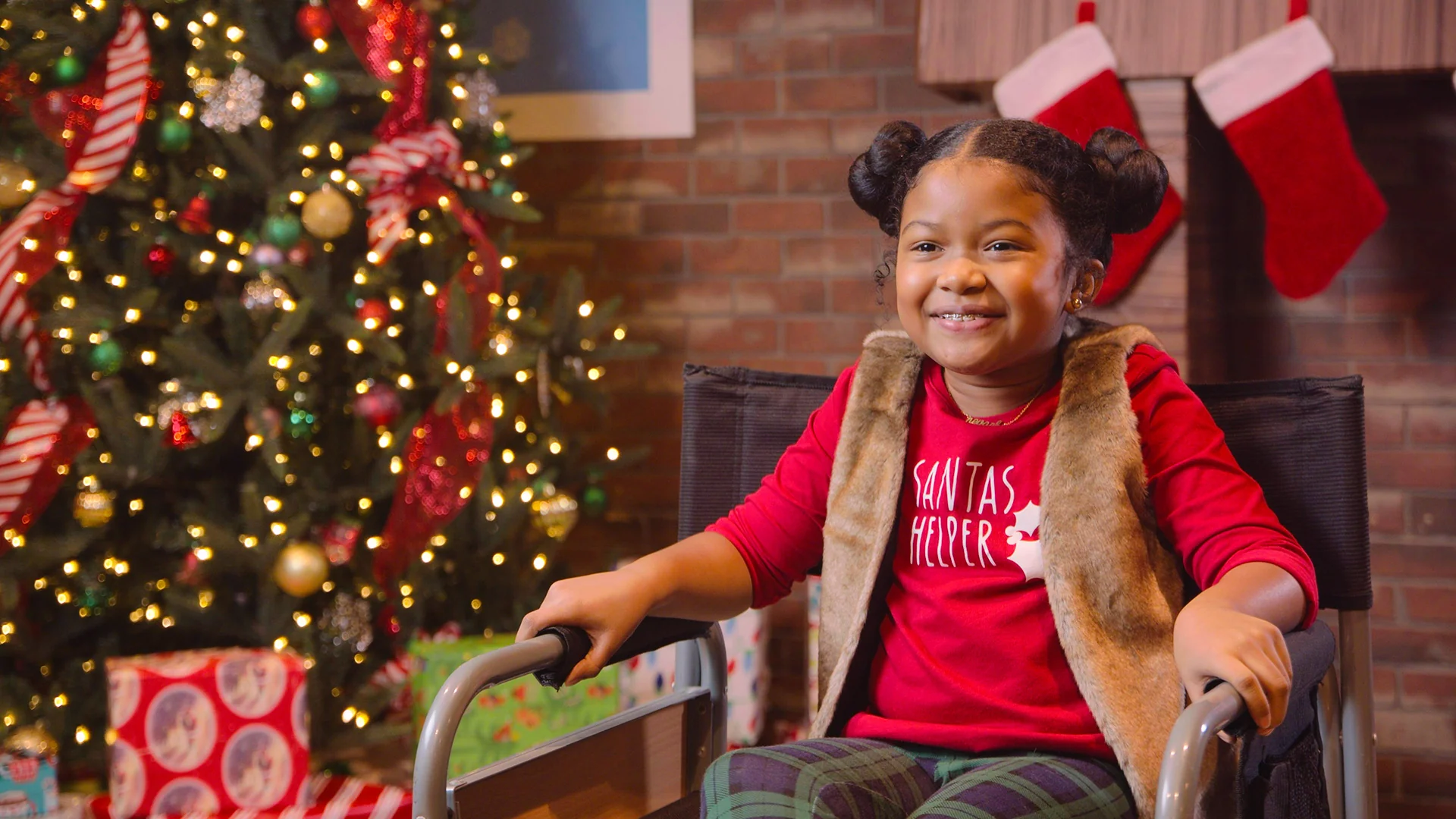 Smiling child in festive attire sitting near a Christmas tree with gifts, celebrating holiday cheer.
