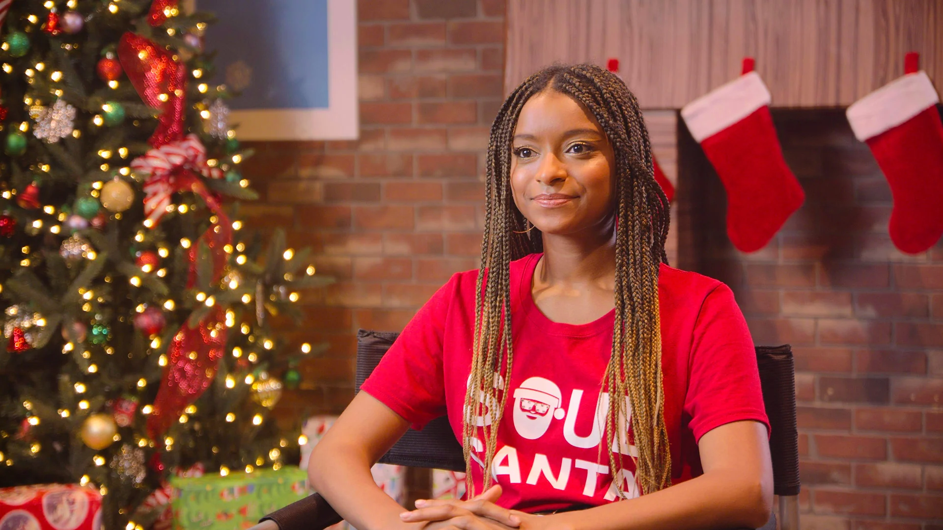 Smiling girl sitting by a decorated Christmas tree, wearing a red shirt with a festive design.