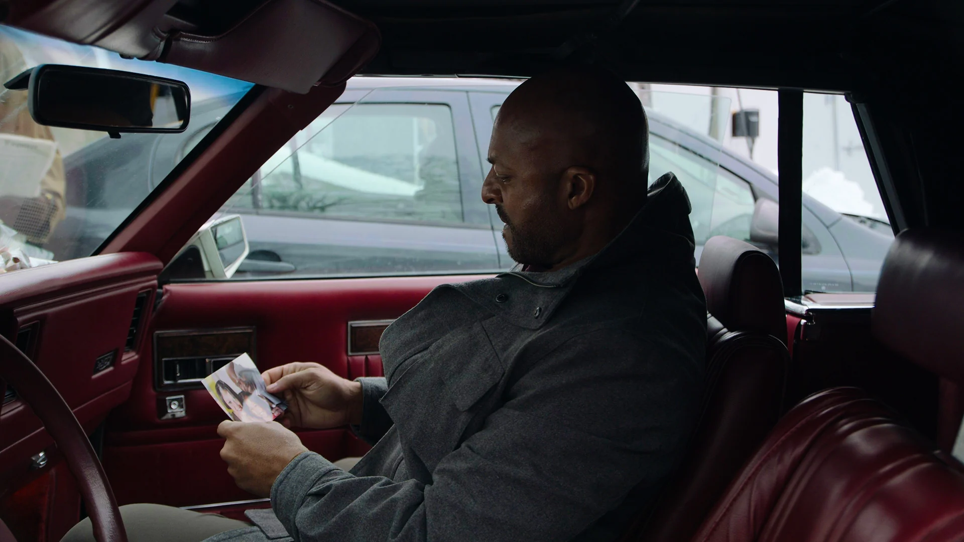 Man sitting in a car, looking at a photo, with a red interior and another vehicle behind.