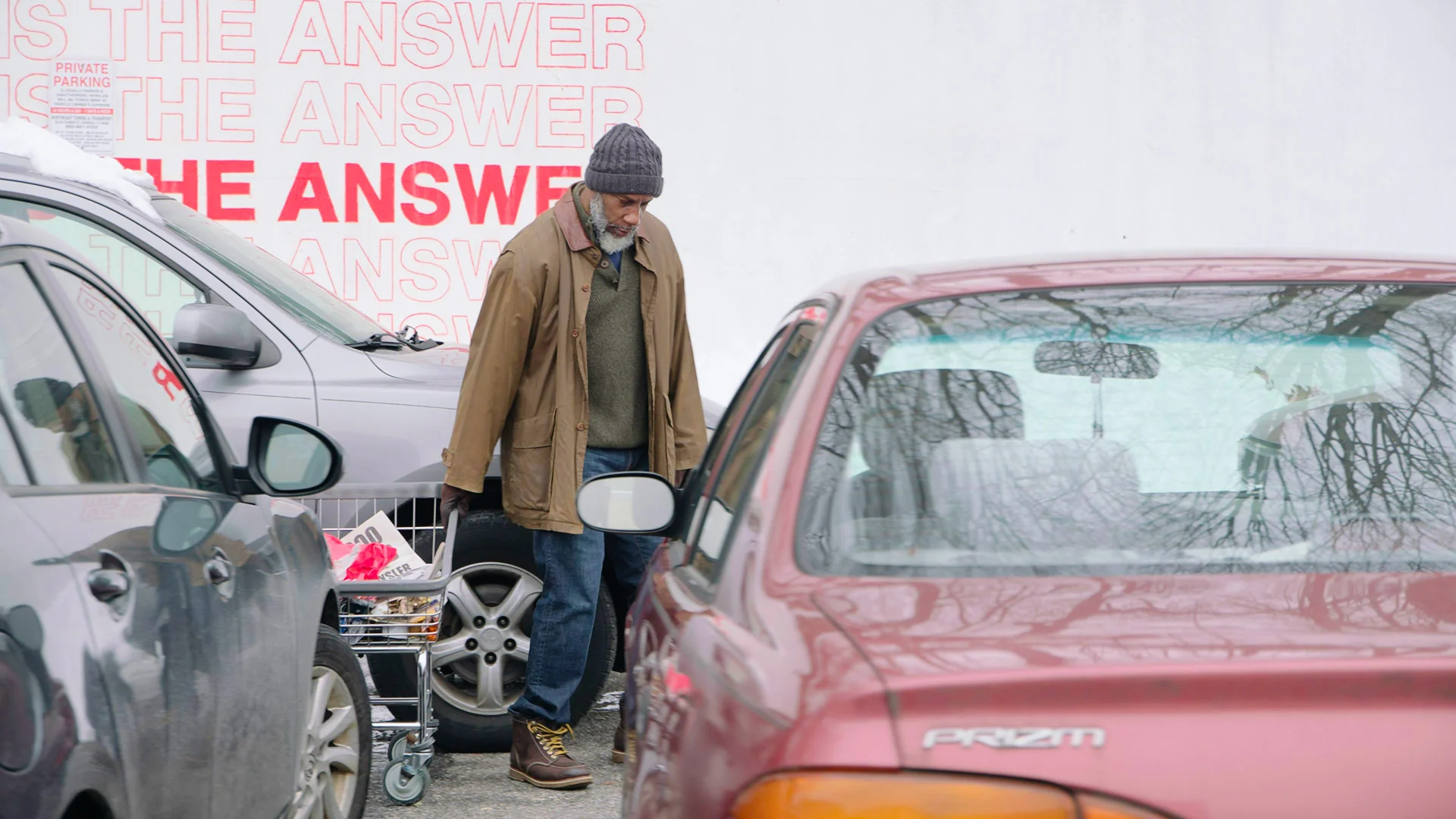 A man with a shopping cart walks between parked cars near a wall with a bold graphic text.