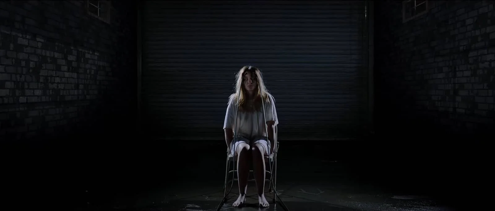 A woman, with cables attached to her, sitting on a chair in a dimly lit, shadowy room.