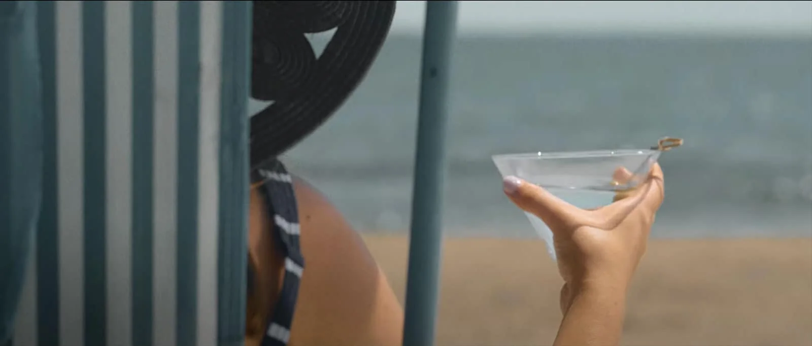 Person holding a drink under a beach umbrella by the ocean.
