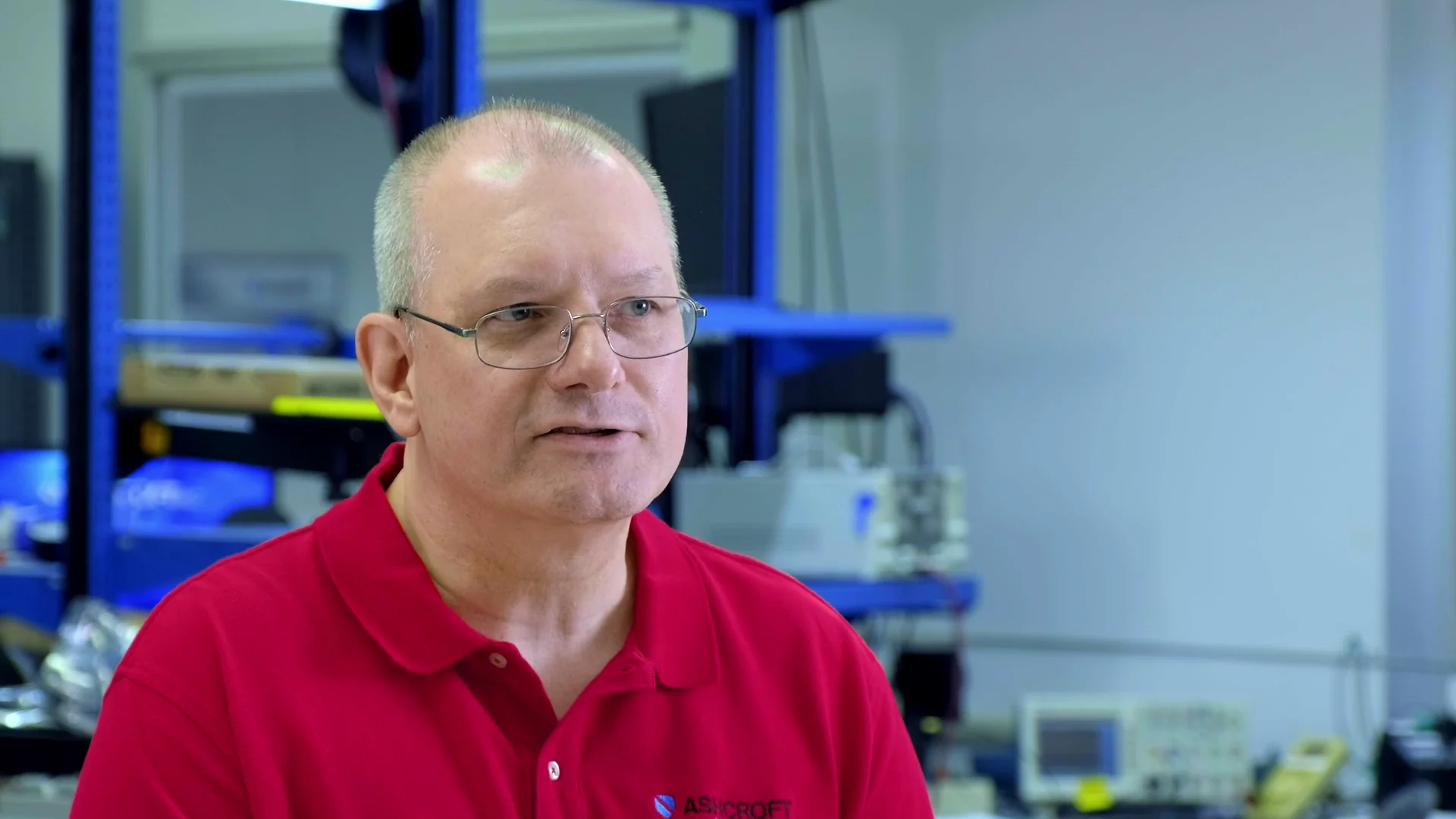 Man in red polo shirt speaking in a technology lab with equipment in the background.