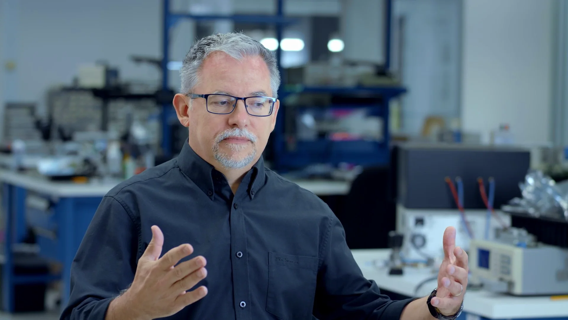 Man in a lab discussing technical concepts with equipment in the background.