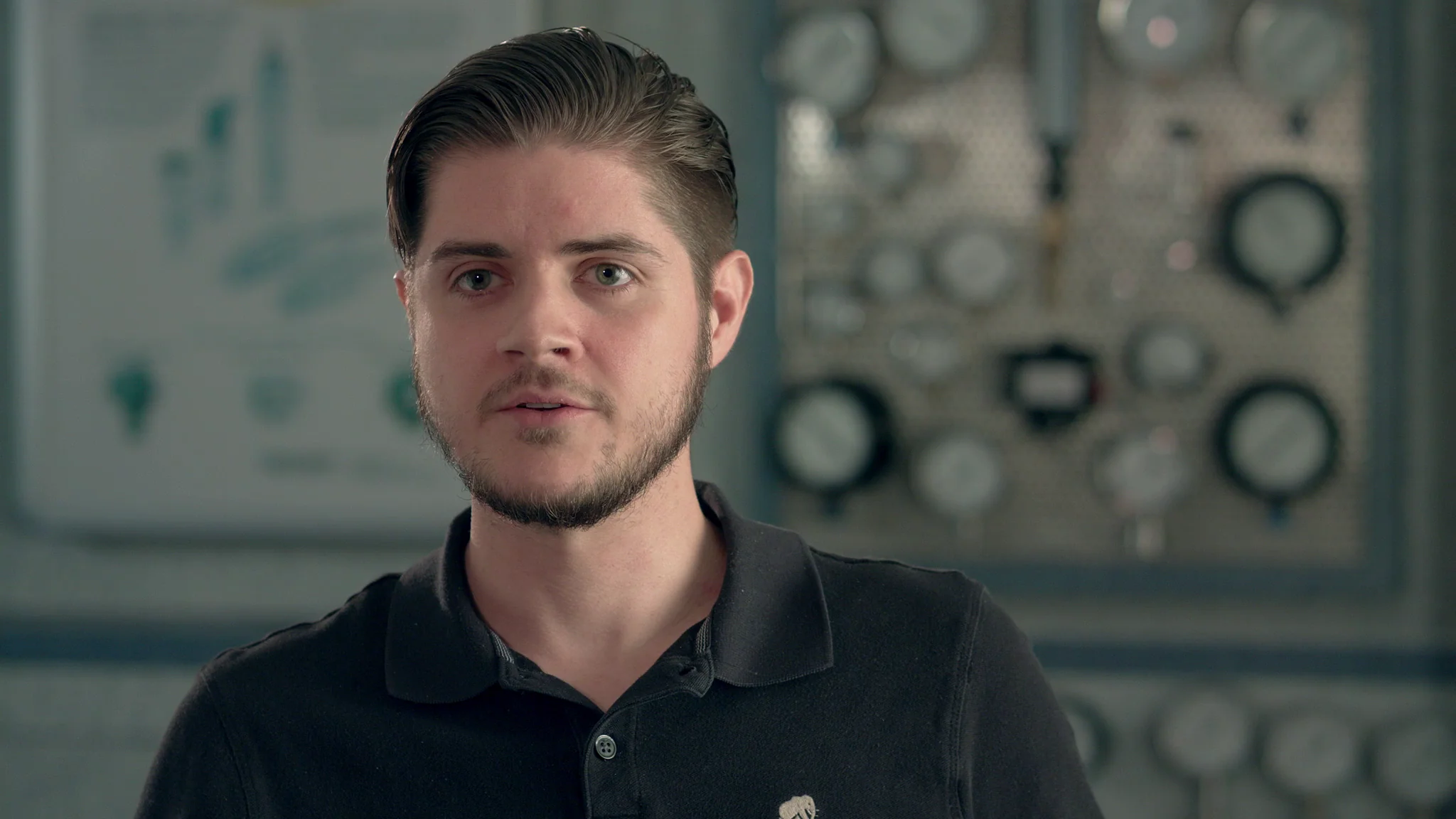 Man in black polo shirt in front of machinery, explaining technical concepts with focused expression.
