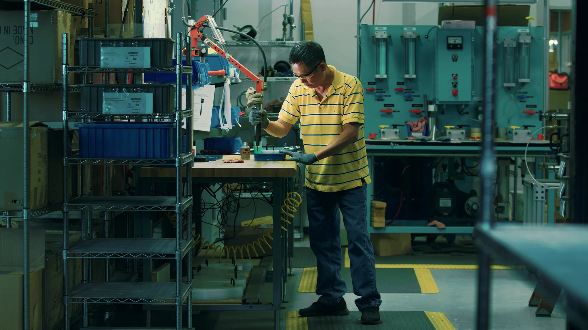 Worker in a workshop using tools, surrounded by shelves and machinery in a production environment.