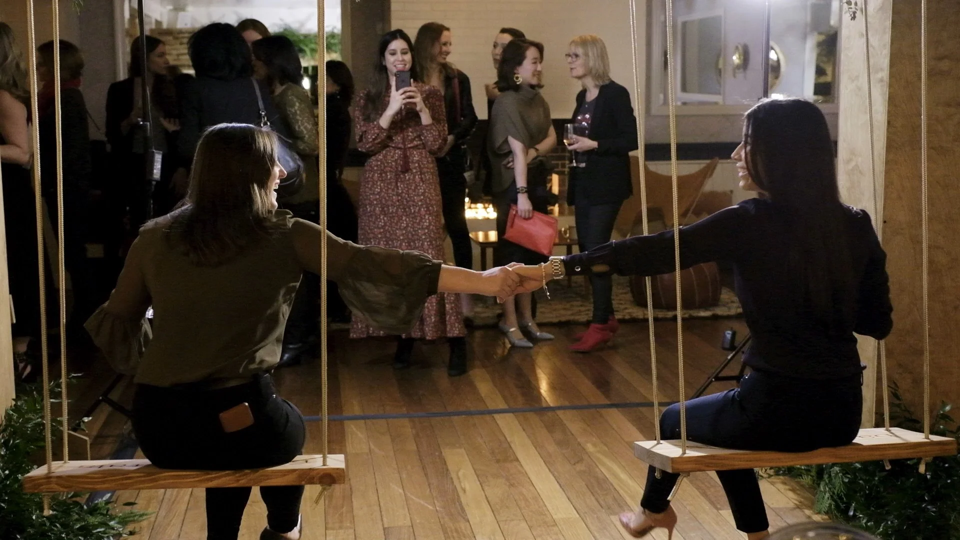 Two women hold hands while sitting on swings at a social gathering.
