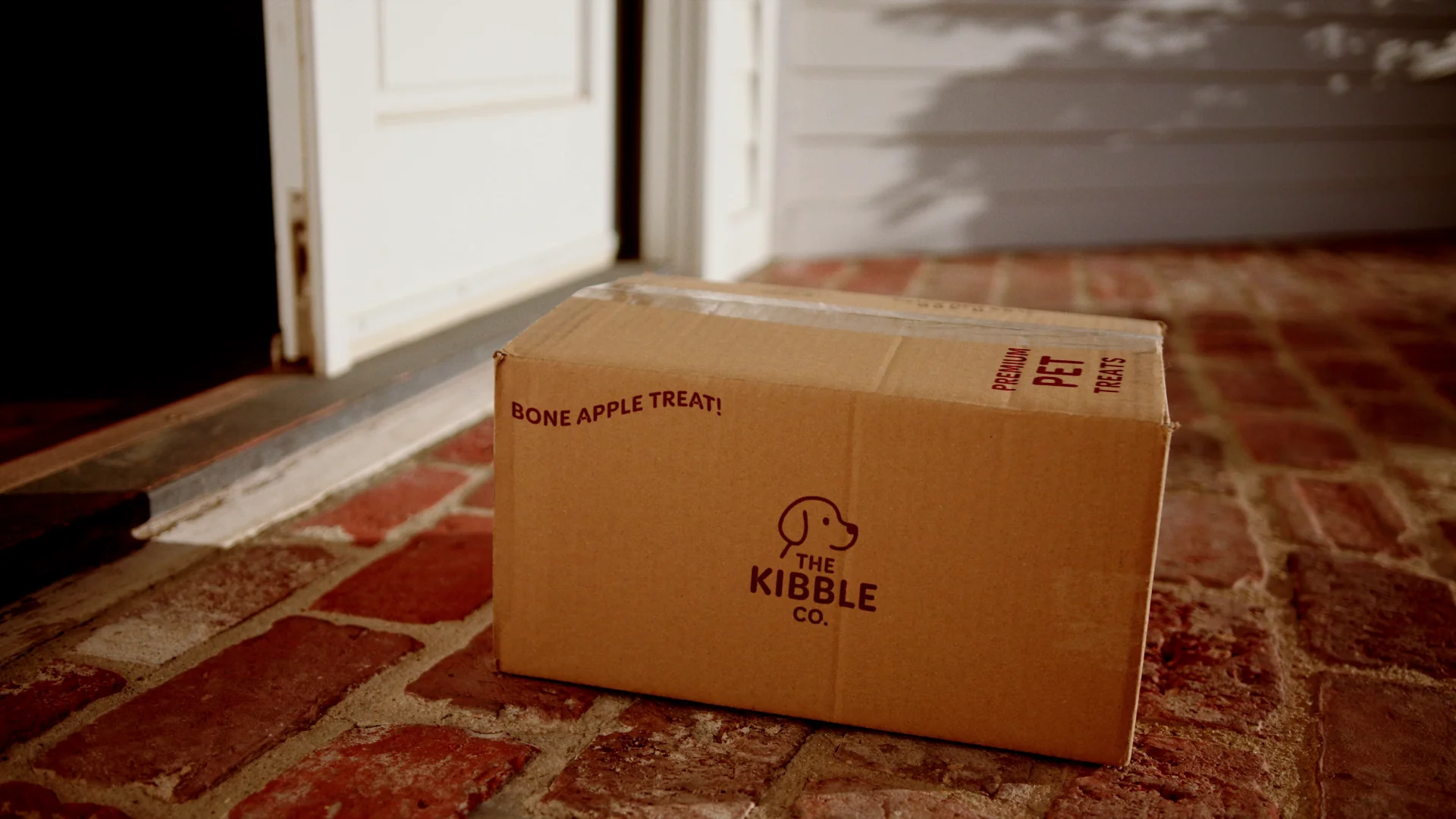 Cardboard box from The Kibble Co. with pet treats, resting on a brick porch.